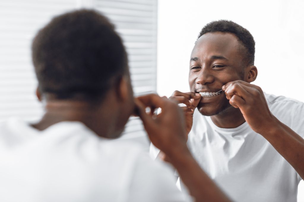 Smiling man flossing his teeth in front of a bathroom mirror, demonstrating good oral hygiene practices, dental care, and the importance of daily flossing for healthy gums.